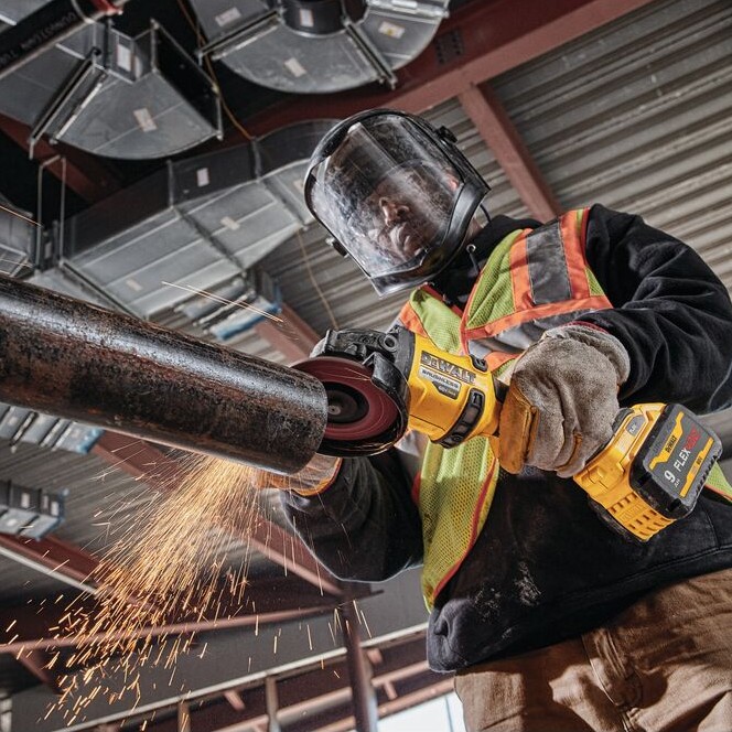 Worker wearing face shield and high-visibility vest using DEWALT cordless angle grinder to grind steel pipe, sparks visible, interior construction site with exposed ductwork overhead.