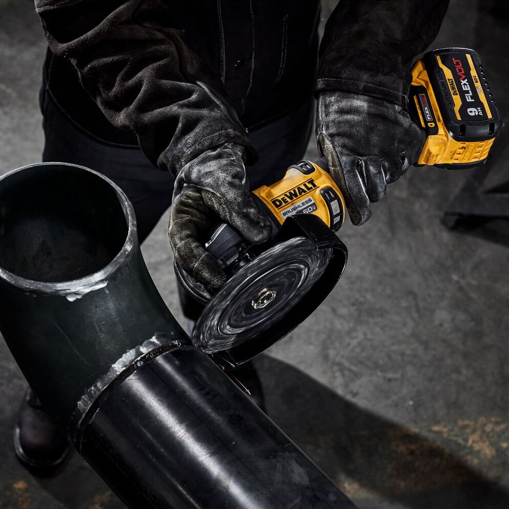 Person wearing black gloves using DEWALT DCG440 60V MAX cordless grinder with 9.0Ah FLEXVOLT battery to grind metal pipe, grinding wheel in contact with pipe edge