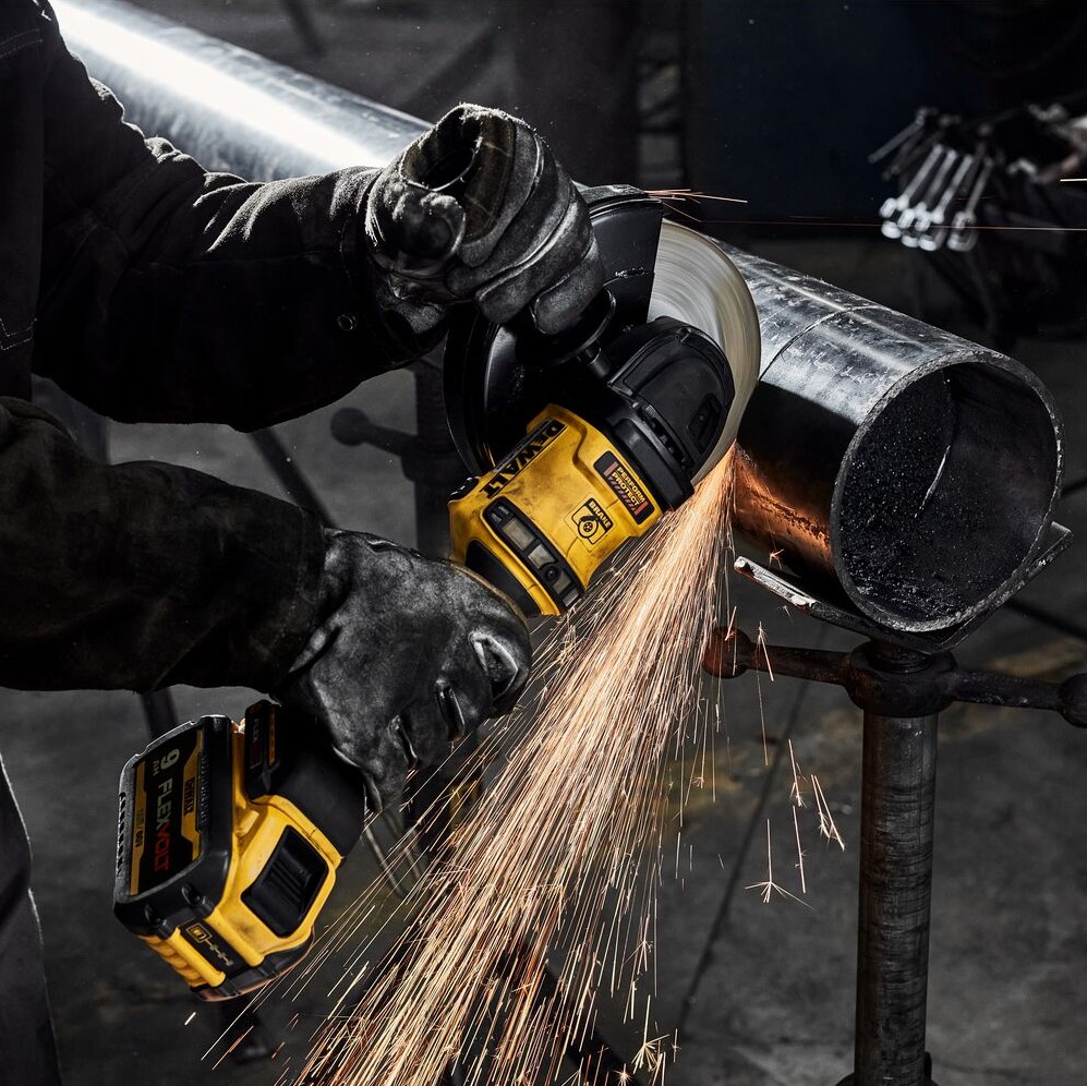 Person wearing black gloves using DEWALT DCG440 60V MAX cordless grinder with 9.0Ah FLEXVOLT battery to cut steel pipe on stand, grinding wheel contacting pipe and sparks flying downward