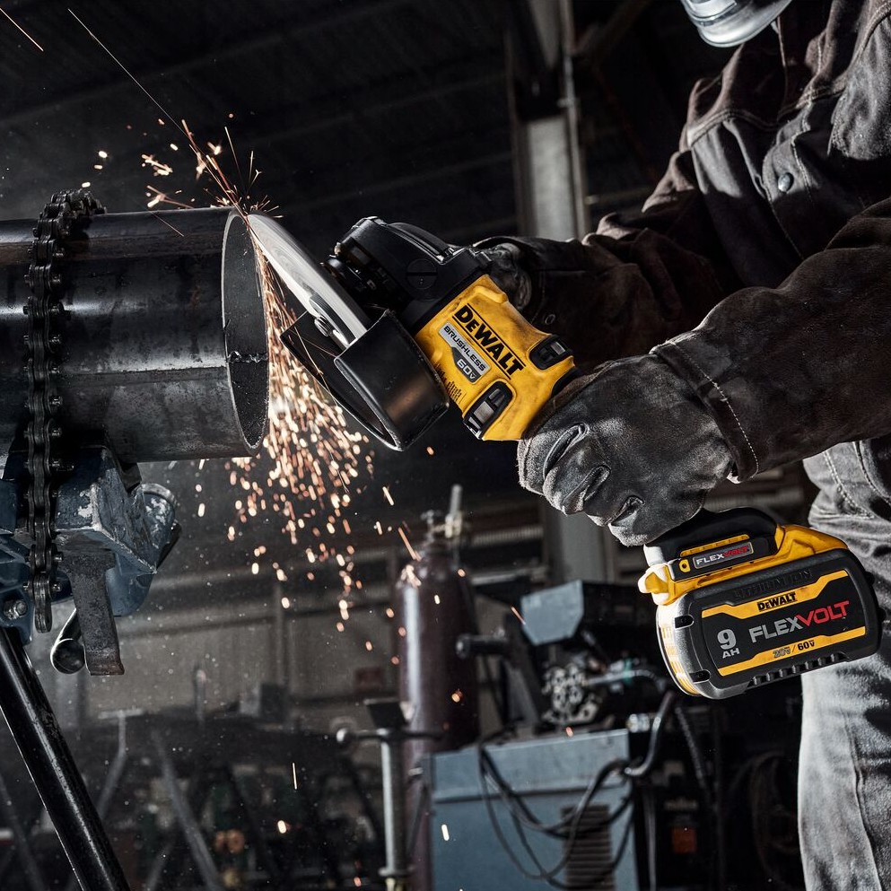 Person wearing gloves using DEWALT DCG440 60V MAX cordless grinder with 9.0Ah FLEXVOLT battery to cut large steel pipe clamped in stand, cutting wheel contacting pipe and sparks flying outward