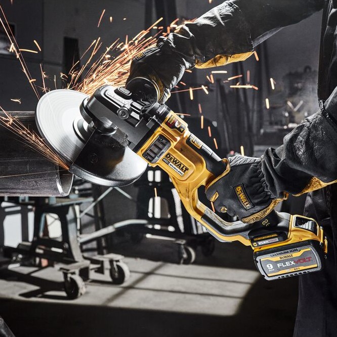 DEWALT 60V MAX FLEXVOLT large angle grinder cutting metal with a cutting wheel, producing sparks, cordless battery installed, two-handed operation with gloves, and steel fabrication work inside an industrial shop.