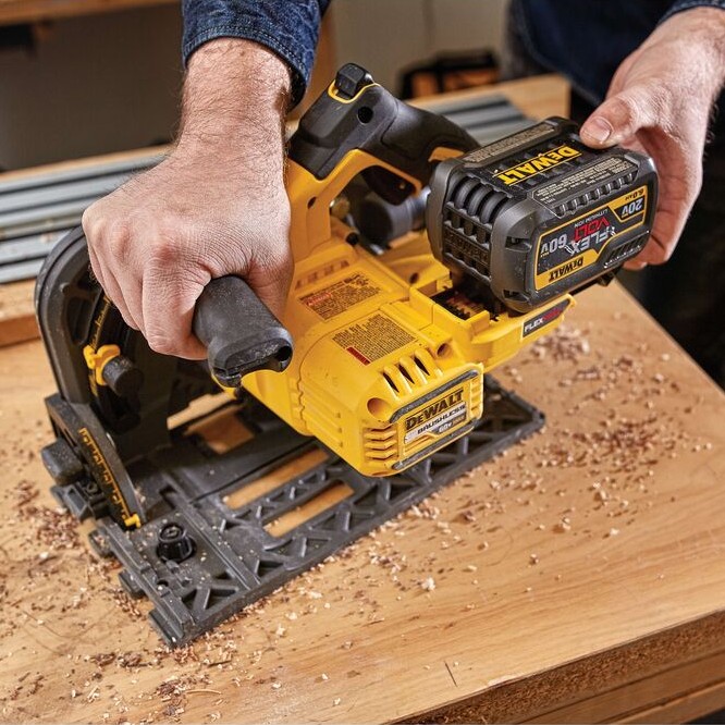 Hands installing a DEWALT FLEXVOLT battery onto a DEWALT 60V MAX brushless 6-1/2 in. TrackSaw resting on a wood work surface, with wood shavings visible nearby.
