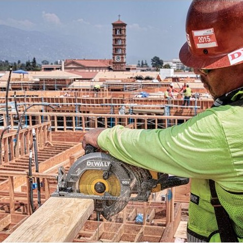 DEWALT FLEXVOLT 60V MAX rear handle worm drive style circular saw cutting dimensional lumber on elevated framed construction site, worker wearing hard hat and safety vest, multi-story wood framing and tower visible in background