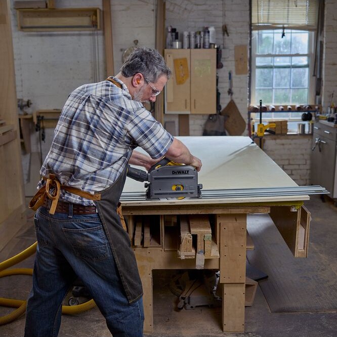 Person using a DEWALT TrackSaw on an aluminum guide rail to cut a large panel on a workbench inside a workshop, with a dust hose connected and tools visible in the background.