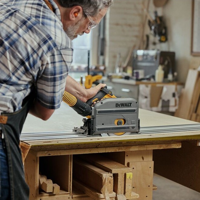 Person wearing safety glasses using a DEWALT TrackSaw on an aluminum guide rail to cut a large panel on a workbench, with a dust extraction hose connected in a workshop setting.