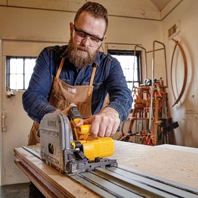 Person wearing safety glasses and a work apron operating a DEWALT TrackSaw on an aluminum guide rail, cutting a wood panel on a workbench inside a workshop.