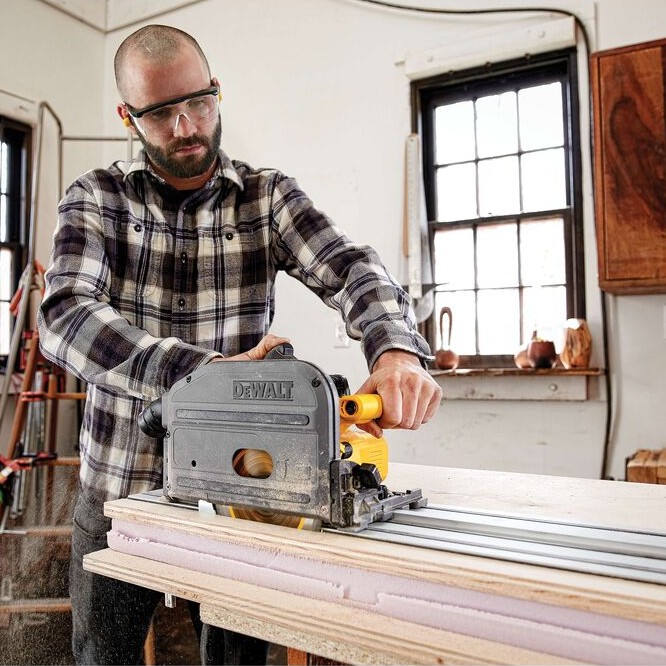 Person wearing safety glasses operating a DEWALT TrackSaw on an aluminum guide rail, cutting a wood panel on a workbench inside a workshop.