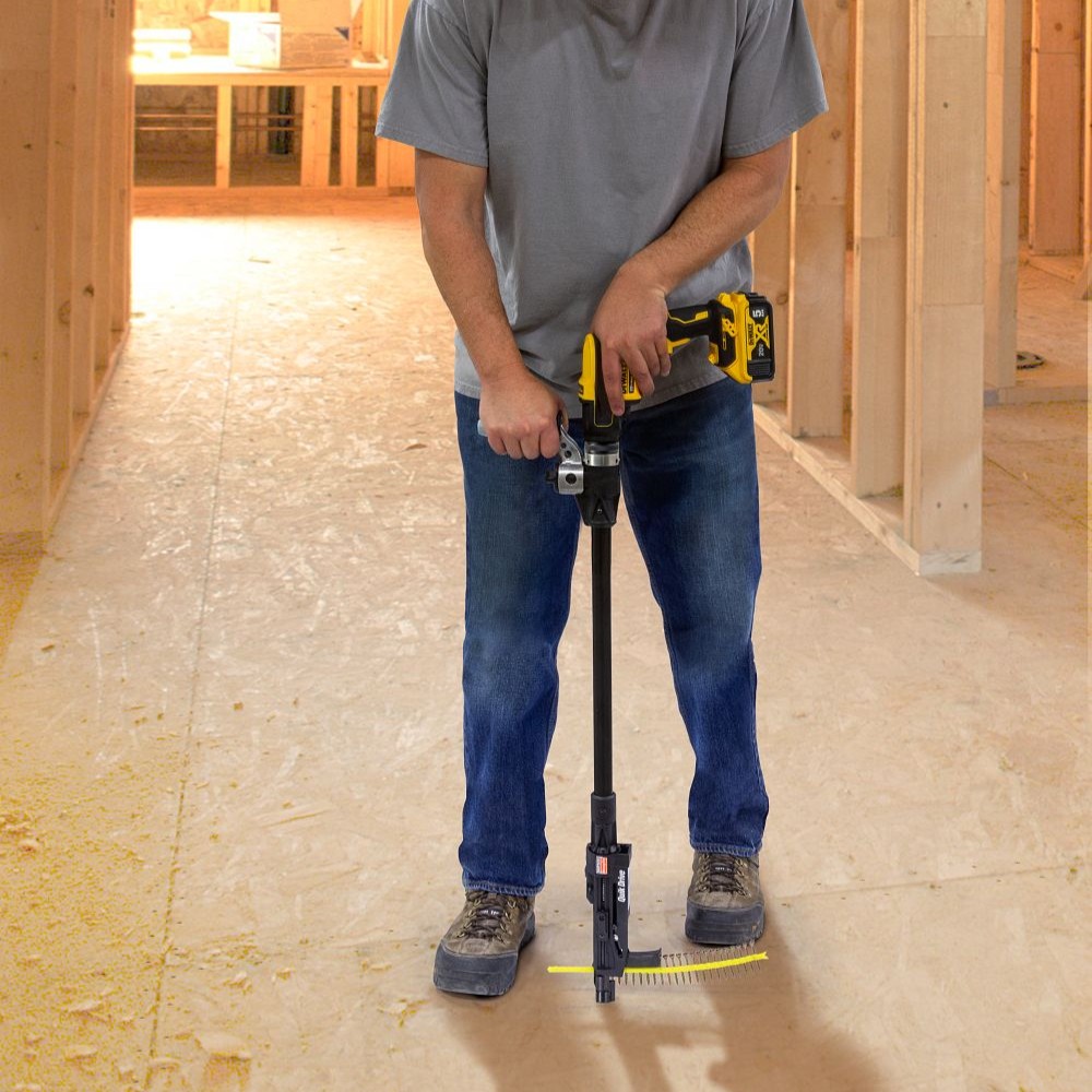 Person standing indoors on a wood subfloor using a Quik Drive PRO200SG2 auto-feed screw driving system with long extension and DEWALT 20V cordless driver, feeding collated screws into the floor.