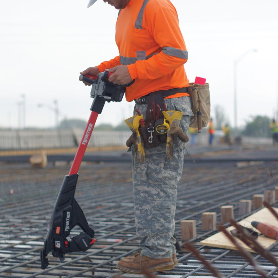 Construction worker wearing high-visibility orange shirt using MAX TwinTier stand-up rebar tying tool while standing on rebar grid at jobsite