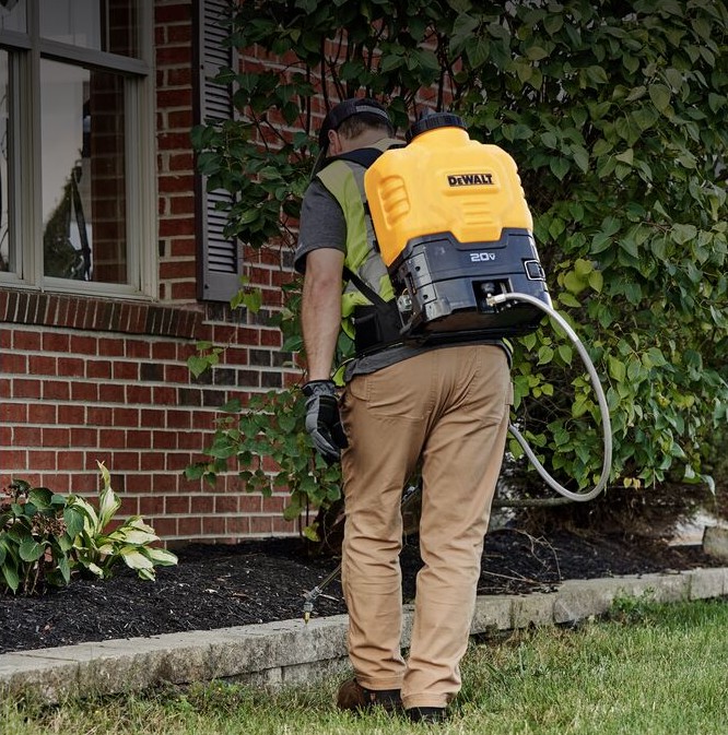 DEWALT 184766 sprayer wand and nozzle kit in use with 20V backpack sprayer, operator spraying along brick house foundation and garden bed, hose and wand visible, outdoor landscaping application image