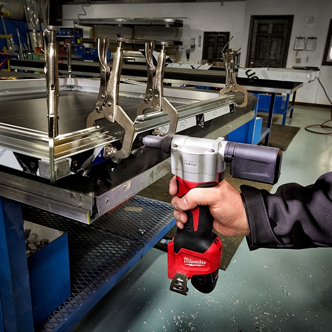Milwaukee 2550-20 M12 rivet tool in use fastening metal frame assembly on workbench with clamps, handheld application close-up in workshop setting