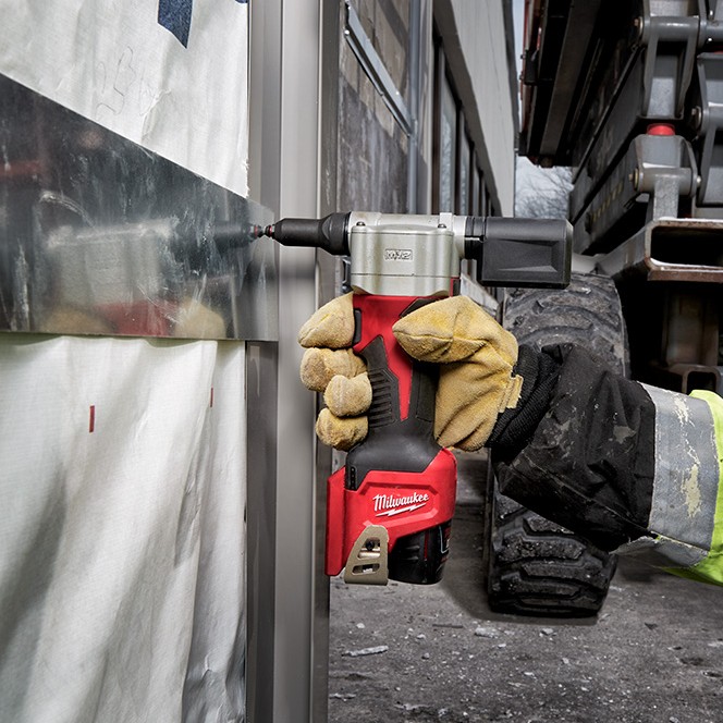 Milwaukee 2550-20 M12 rivet tool installing rivet into metal siding panel, gloved hand holding tool, close-up application on exterior construction surface