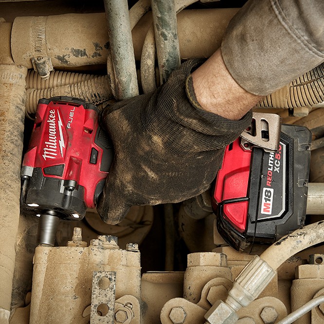 Milwaukee 2855-22R M18 FUEL 1/2" compact impact wrench in use inside heavy equipment engine bay, gloved hand removing fastener in tight space with XC5.0 battery installed