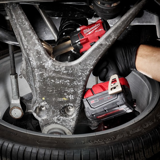 Milwaukee 2855-22R M18 FUEL 1/2" compact impact wrench removing suspension fastener inside wheel well, gloved hand using tool in tight automotive space with XC5.0 battery attached