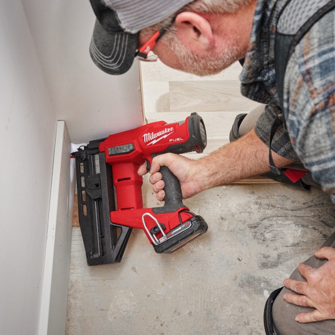 Milwaukee 3020-20 M18 FUEL 16 gauge straight finish nailer installing baseboard trim in corner, application shot showing nose placement tight to wall and compact body for confined space work