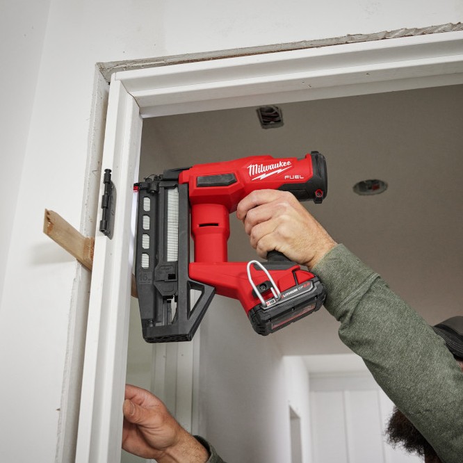 Milwaukee 3020-20 M18 FUEL 16 gauge straight finish nailer installing trim on door frame, angled application shot showing nailer nose placement and compact body in tight space