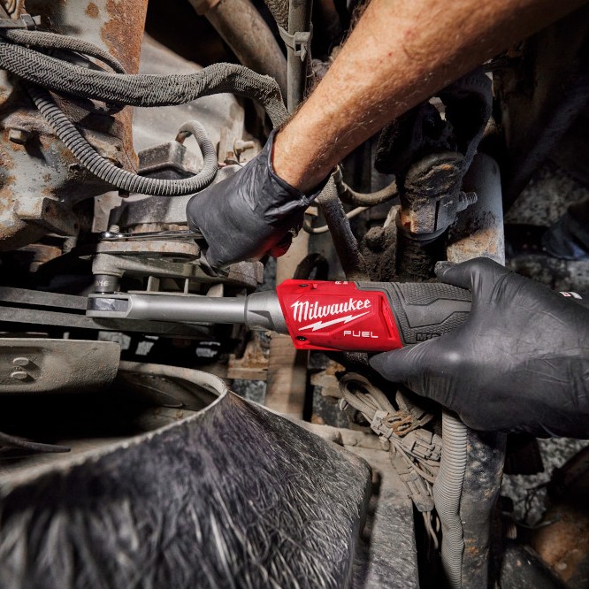 Milwaukee M12 FUEL INSIDER extended reach box ratchet in use on vehicle undercarriage, gloved technician loosening fastener in tight suspension area with long neck access