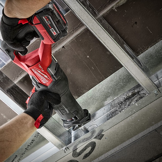Milwaukee M18 FUEL SAWZALL reciprocating saw cutting steel framing, operator wearing gloves using cordless saw with battery attached, blade engaged on metal stud channel in construction setting