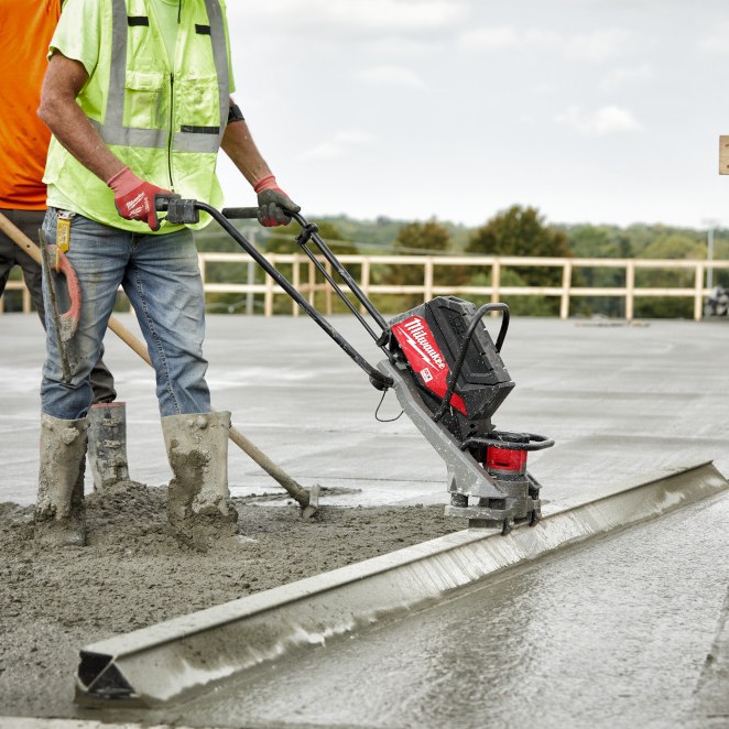 Milwaukee MXF381-2CP MX Fuel vibratory screed in use finishing wet concrete slab, operator guiding screed bar along slab edge with fresh concrete surface and railing in background