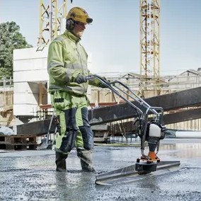 Husqvarna 967858901 10 ft screed blade in use on BV30 power screed, operator leveling wet concrete slab, construction site background with worker and rebar structure visible