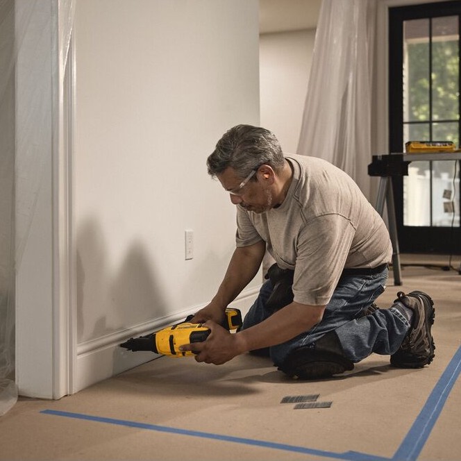 DEWALT DCN650B 20V MAX XR 15 gauge angled finish nailer in use installing baseboard trim, contractor kneeling on floor fastening molding along wall, cordless nailer with angled magazine and battery attached in real jobsite setting