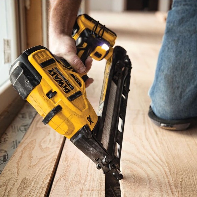 DEWALT DCN650B 20V MAX XR 15 gauge angled finish nailer in use installing hardwood flooring, close-up of cordless nailer driving angled finish nails along floor edge with LED work light illuminated and battery attached
