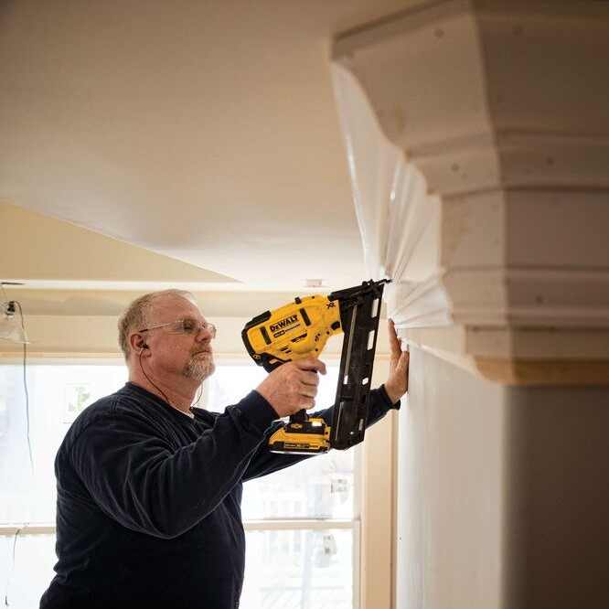 DEWALT DCN650B 20V MAX XR 15 gauge angled finish nailer in use installing crown molding, contractor fastening trim near ceiling with cordless angled nailer, battery attached in interior finish carpentry setting