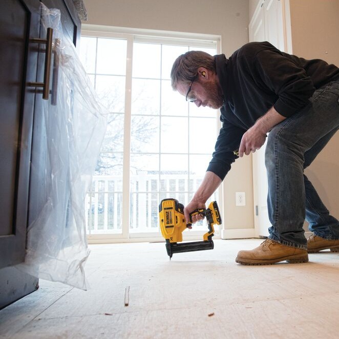 DEWALT DCN681D1 20V MAX XR 18GA narrow crown stapler in use installing flooring underlayment, worker kneeling and fastening wood subfloor near patio door in residential jobsite setting