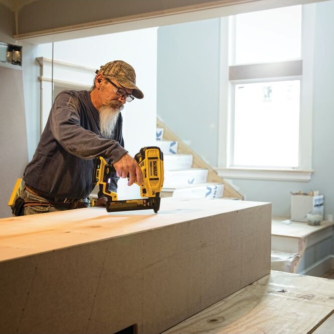 DEWALT DCN681D1 20V MAX XR 18GA narrow crown stapler in use fastening wood trim on interior jobsite, worker using cordless stapler on large wood assembly near staircase in residential setting