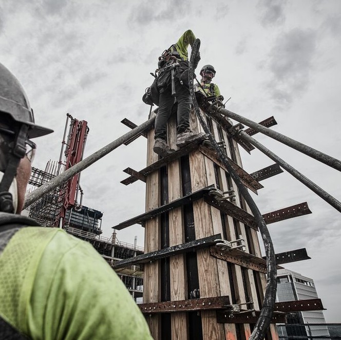 DEWALT DCPS320AG2 POWERSHIFT concrete vibrator used for vertical column pour, workers consolidating concrete at height with hose routed up formwork and rebar structure visible