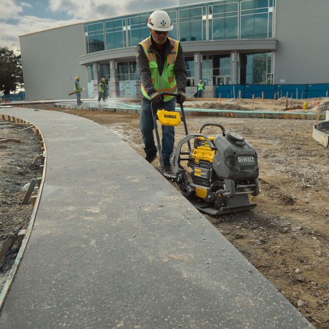 DEWALT POWERSHIFT forward plate compactor compacting soil along concrete sidewalk edge at commercial building jobsite, operator in hard hat and high-visibility vest guiding unit beside formed curb