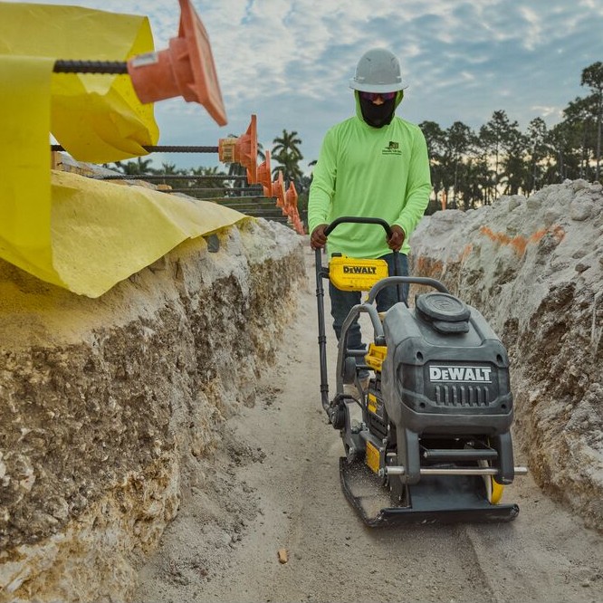 DEWALT POWERSHIFT forward plate compactor compacting soil in trench, operator wearing hard hat and face covering guiding unit along excavation with safety caps and rebar visible on left side