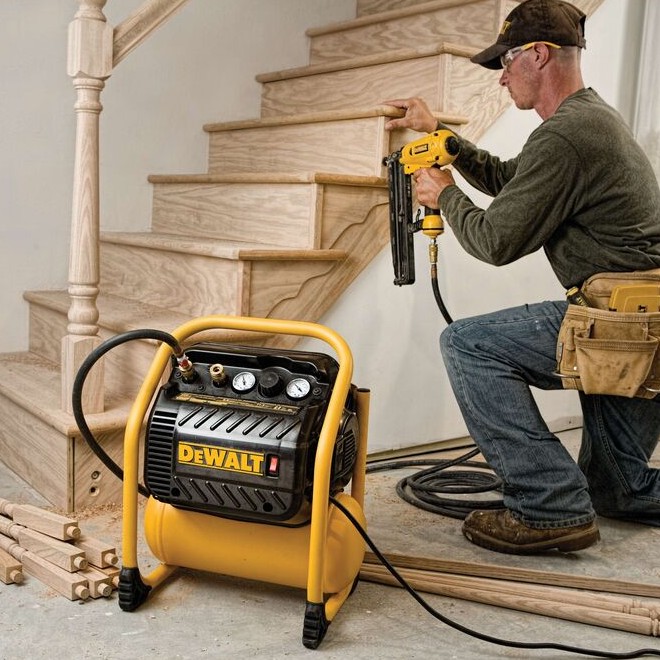 DEWALT DWFP55130 200 PSI quiet trim compressor powering finish nailer during stair trim installation, contractor installing wood stair nosing, air hose connected, indoor carpentry jobsite scene