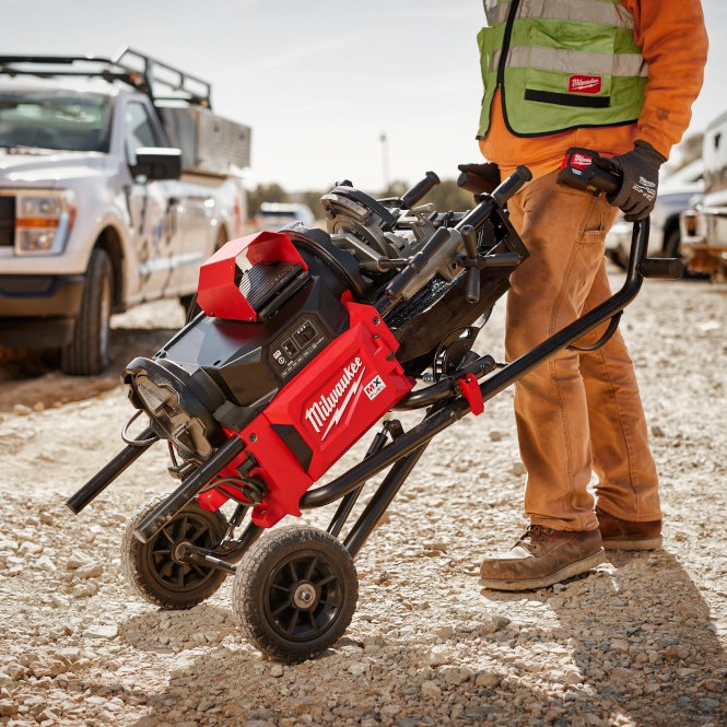 Milwaukee MXF512-2XC MX FUEL pipe threading machine on transport cart being wheeled across gravel jobsite, worker holding handles, machine tilted on large wheels, service truck in background