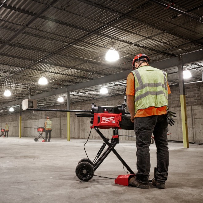 Milwaukee MXF512-2XC MX FUEL pipe threading machine set up indoors on jobsite floor, worker operating machine with long pipe, large open industrial space with multiple workers and machines in background