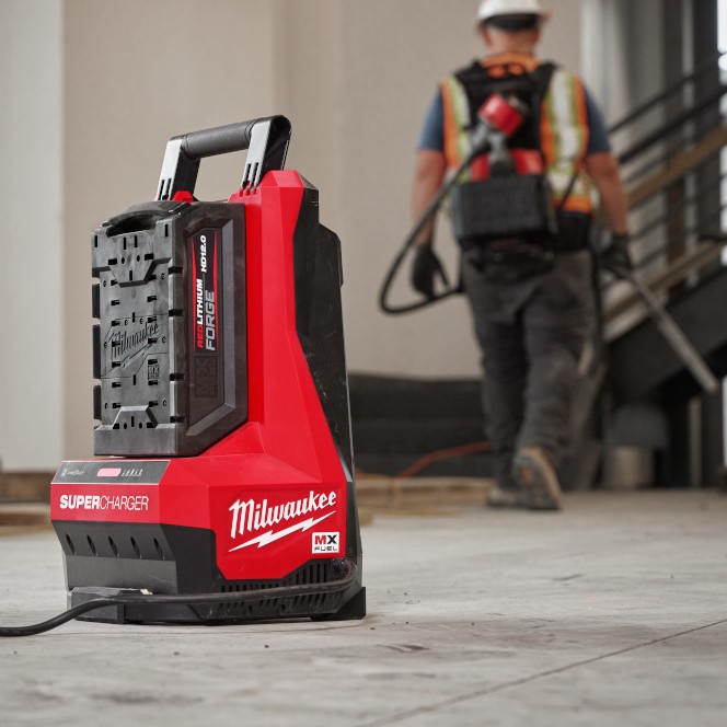 Milwaukee MXFSC MX FUEL Super Charger with REDLITHIUM FORGE battery installed on floor, power cord plugged in, construction worker walking in background wearing safety gear