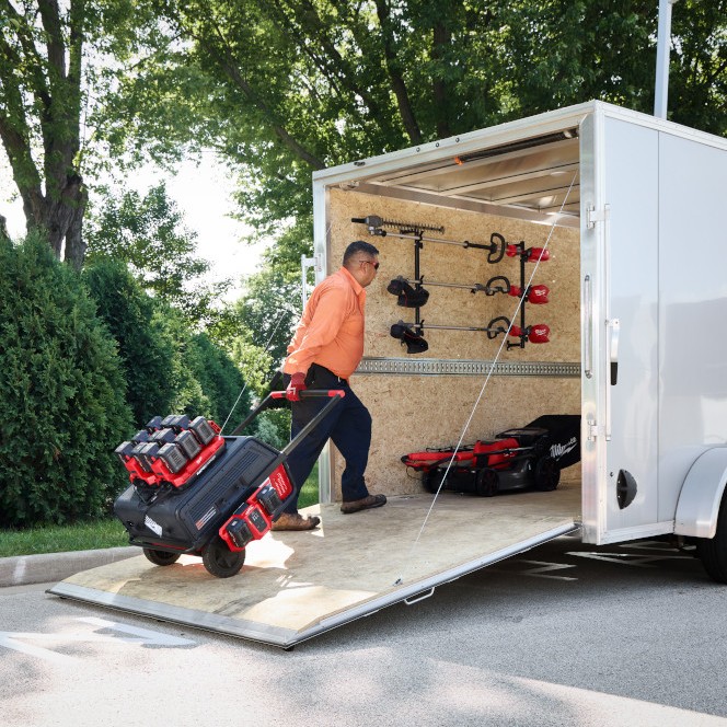 Worker transporting a Milwaukee ROLL-ON™ power supply loaded with M18™ battery chargers and batteries into an enclosed trailer, demonstrating portable jobsite power and equipment transport with all-terrain wheels.