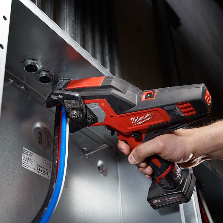MILWAUKEE M12 600 MCM cable cutter cutting large electrical cable inside a crowded panel, showing the compact open-jaw design in use.