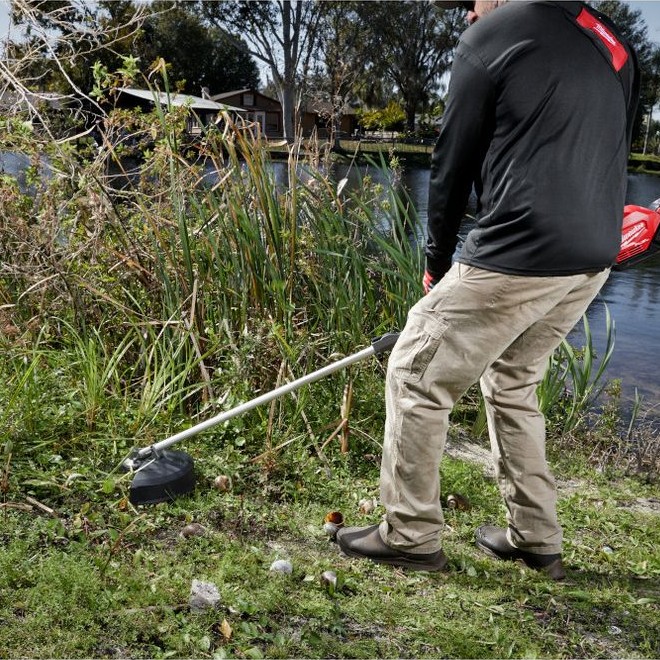 MILWAUKEE M18 FUEL QUIK-LOK Brush Cutter Attachment in use clearing dense weeds and overgrown vegetation along a pond edge.