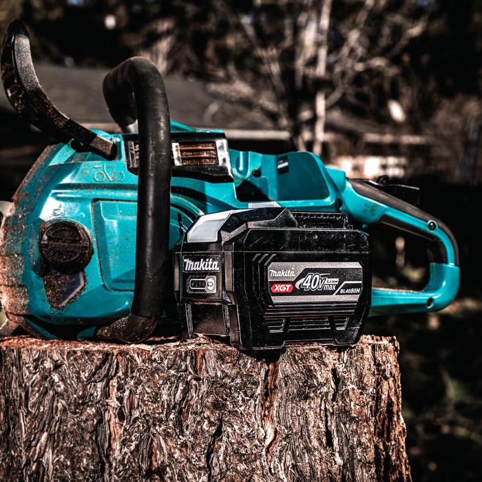 Makita 40Vmax XGT 8.0Ah High Power Battery beside a cordless chainsaw on a tree stump at an outdoor worksite.